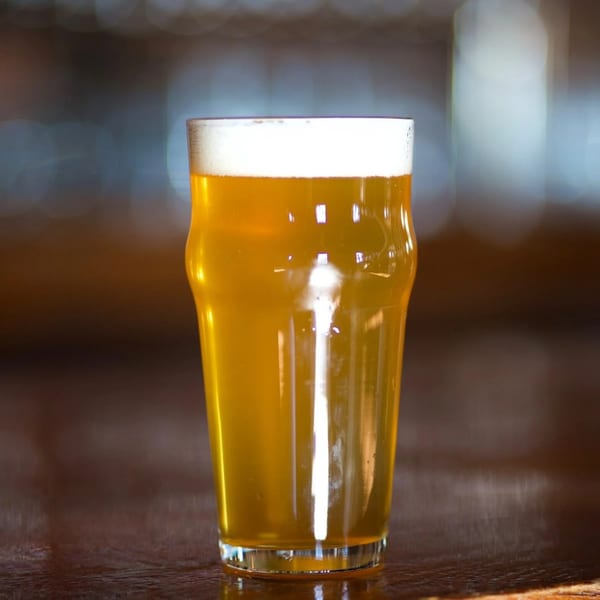 A pint glass of golden beer with a foamy head sits on a dark wooden surface.