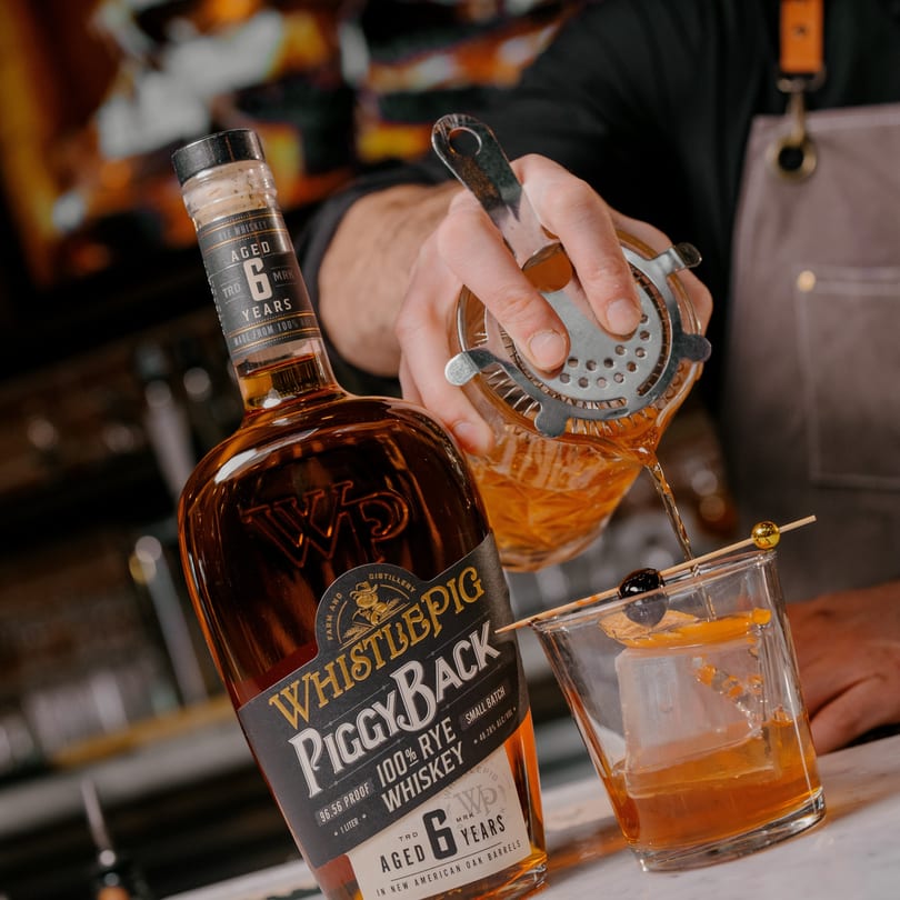 A bartender pours WhistlePig PiggyBack 100% Rye Whiskey from a shaker into a glass with ice and garnish.