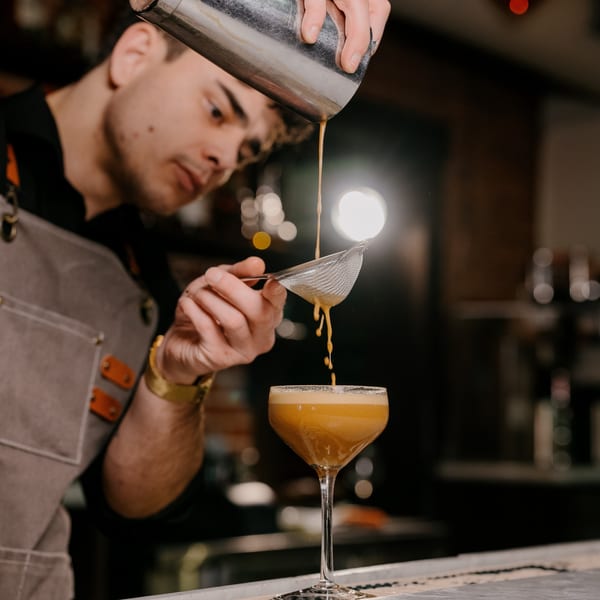 Bartender straining a frothy orange cocktail from a shaker into a coupe glass.