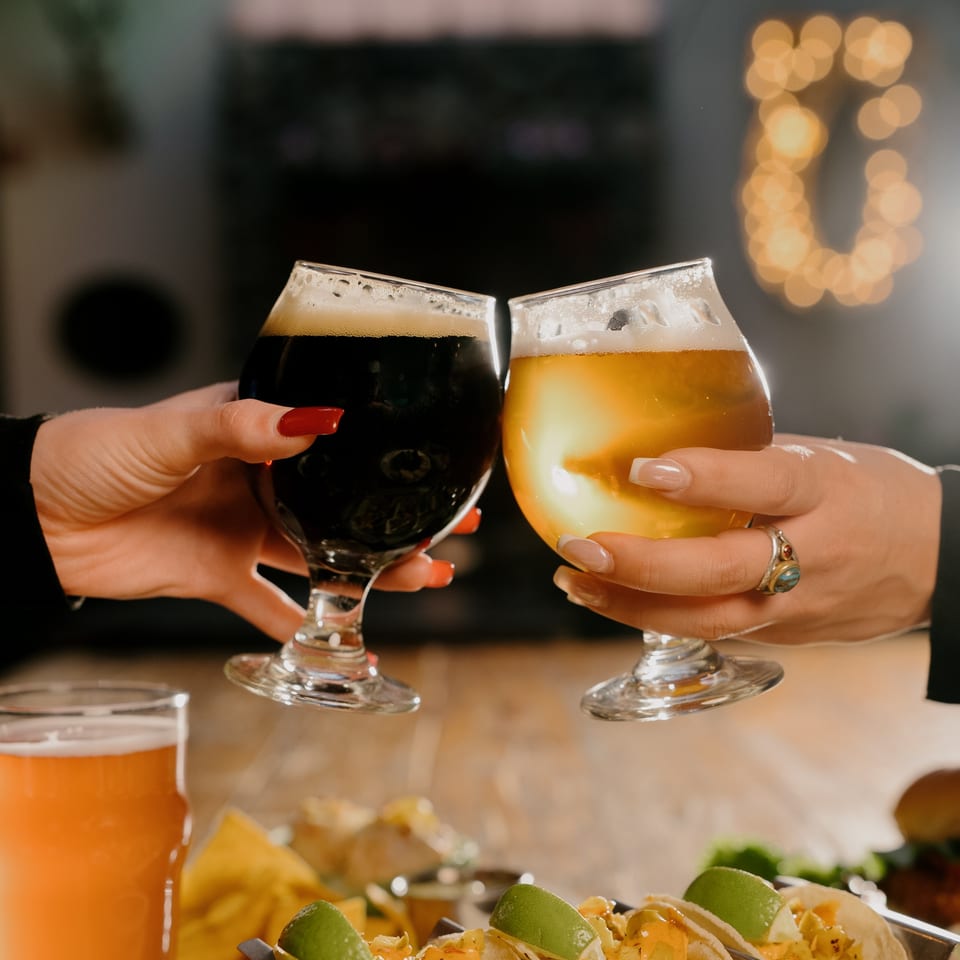 Two people toast dark and light beers over a table of tacos and nachos.