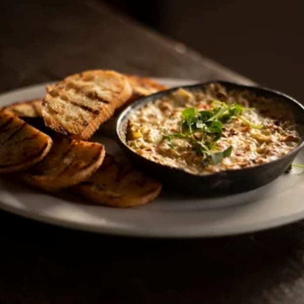 A cast iron skillet of creamy dip topped with greens, served with grilled bread slices.