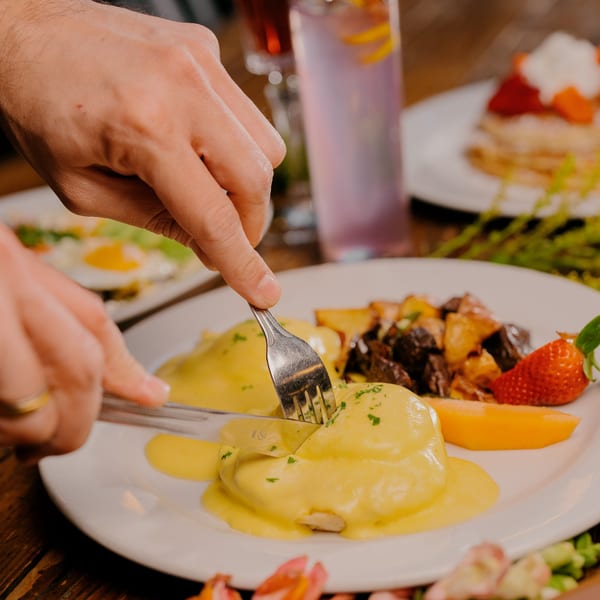 Hands cutting into Eggs Benedict with hollandaise sauce, served with roasted potatoes and fruit.
