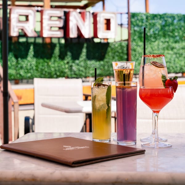 Four colorful cocktails and a menu on a marble table with a "RENO" sign in the background.