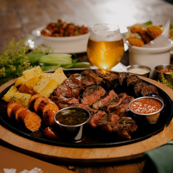 Sizzling platter of grilled steak, sausages, and roasted potatoes with dipping sauces and beer.