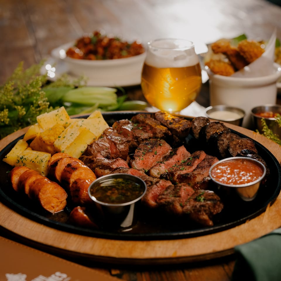 Sizzling platter of grilled steak, sausages, and roasted potatoes with dipping sauces and beer.