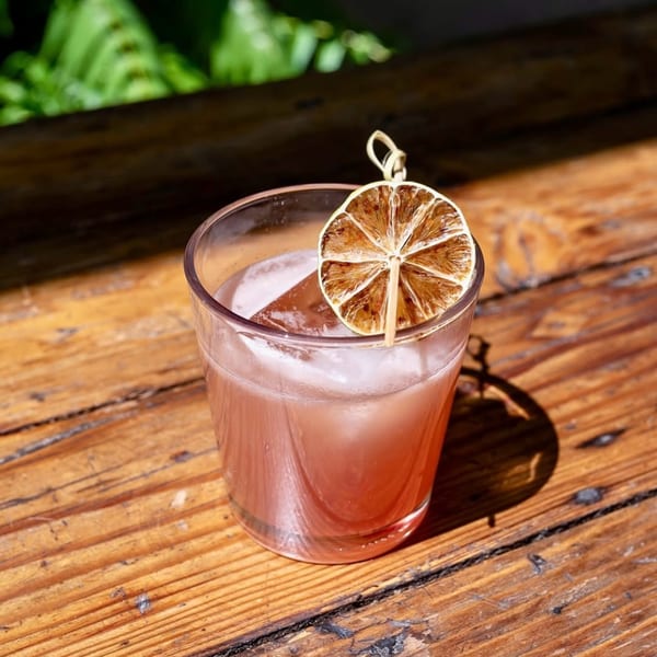 A pink cocktail with a dried citrus slice garnish sits on a rustic wooden table.
