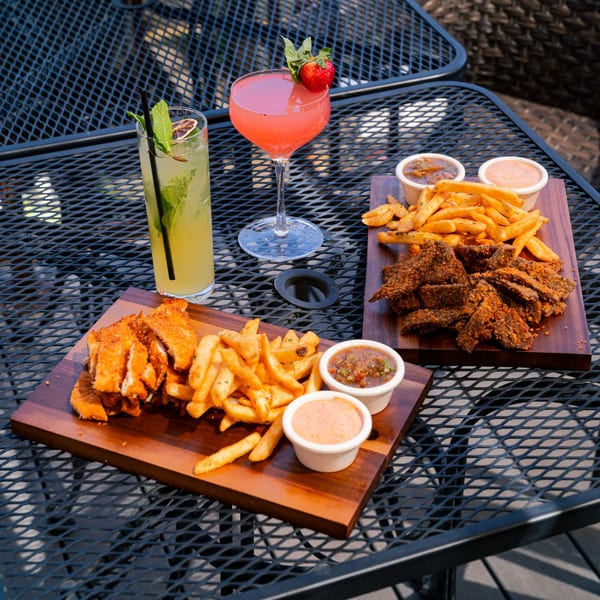 Two cocktails and two wooden boards with fries, sauces, and fried chicken or beef on a patio table.