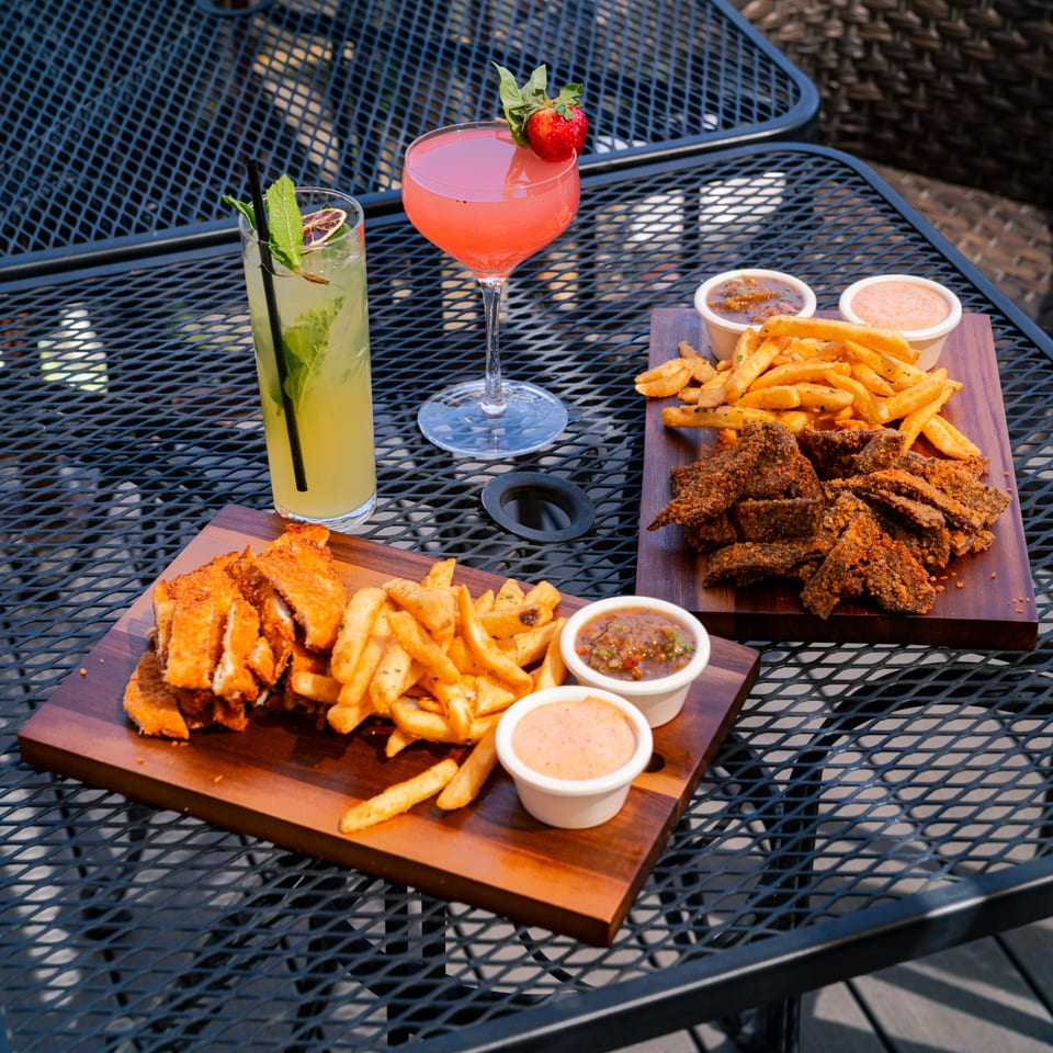 Two cocktails and two wooden boards with fries, sauces, and fried chicken or beef on a patio table.