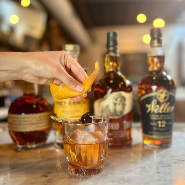Hand garnishing an Old Fashioned cocktail with an orange peel, surrounded by bourbon bottles.